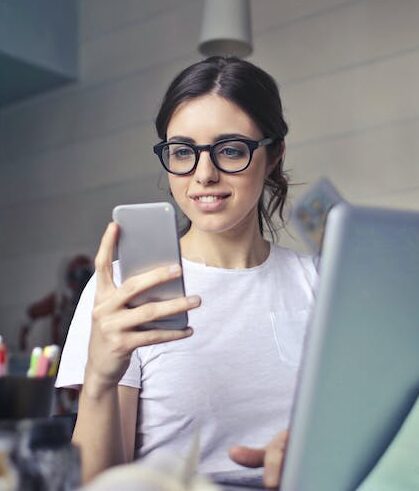 Woman in White T-shirt Holding Smartphone in Front of Laptop